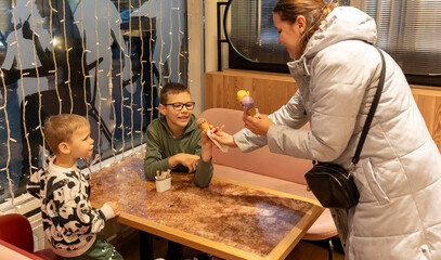 Mother giving ice cream cones to two excited sons sitting at cafe table with festive lights in background. Concept of treating children to dessert, family outing surprise and parenting joyful moments.