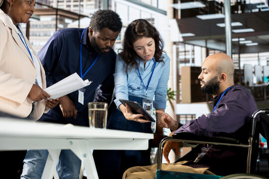 Multiethnic employees team sharing ideas and details for achieving goals, discussing about data analytics and reports. Middle eastern man meeting with colleagues in disability friendly space.