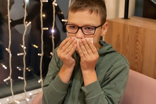 Boy in glasses wiping mouth with napkin after eating at cafe with fairy lights decoration by window on winter evening. Concept of table manners, childhood hygiene habits and family outing moments.