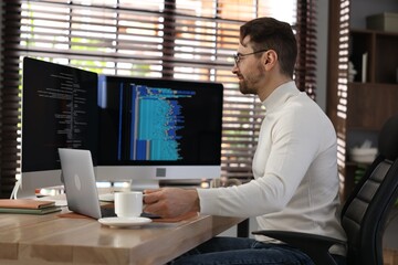 Programmer in glasses working on computer with dual monitors and laptop at table in office