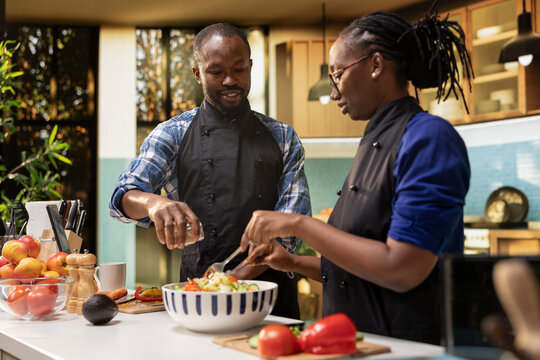 African american man seasoning the salad with mix of spices from a jar, pouring condiments for a tasty vegetarian meal. Smiling woman mixing fresh vegetables together, healthy lifestyle.