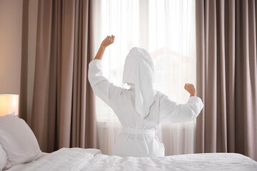 Woman wearing bathrobe with hair wrapped in towel on bed indoors, back view