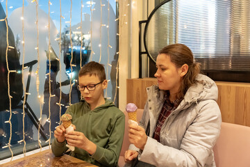 Mother watching son eating ice cream cone with sprinkles at cozy cafe table with fairy lights by window on winter day. Concept of family quality time, sweet indulgence and parent child bonding moments
