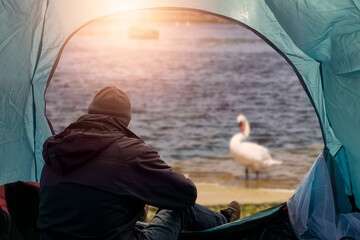 Homeless man sitting in a blue light tent by the entrance and looking at a swan and river, there is warm sun glow in the background and boats. Poverty and social issue. Economy and finance crisis.