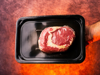 Premium top quality beef rib eye steak in black plastic vacuum packed package in butcher hand, red hot flaming fire background. High value and cost meat product with expensive price.