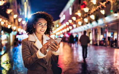 A smiling woman uses her smartphone while walking through a brightly lit street at night, surrounded by festive lights and reflections.