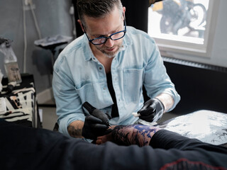 Tattoo artist working on intricate Japanese landscape sleeve with reference stencil visible on table in bright studio lighting.