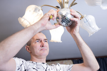 A middle-aged man repairs a chandelier at home. He's focused on his task, changing the light bulbs.