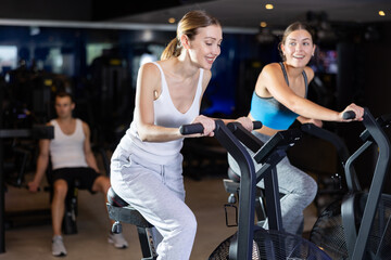During classes on air bike, girls chat sweetly and smile with female friend. Two young women...