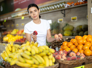 Young woman shopping in vegetable store and buy donut peach. Customer carefully examine goods, thinking about buying. Self-service store, vegetables and fruits from all over world.
