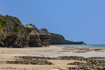 Scenic tropical beach with thatched huts on stilts and exposed coral at low tide.