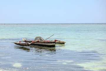 Traditional wooden fishing boat with a trap floats in clear turquoise water, a person walks in the distance.