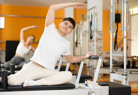 Young guy enjoying Pilates class on reformer in brightly colored studio, doing side bend with arm extended overhead to stretch core muscles, improve posture and flexibility