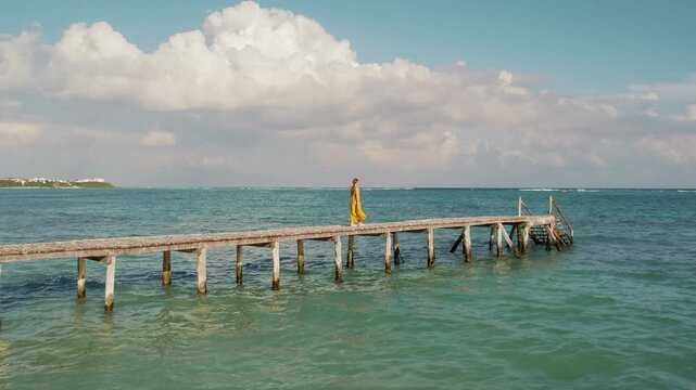 Woman in maxi dress walking alone on a rustic pier over turquoise sea in Mexico