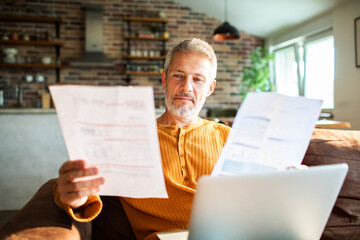 Mature man reviewing bills with laptop at home, focused