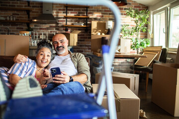 Mature couple happily relaxing with smartphone in new home with moving boxes