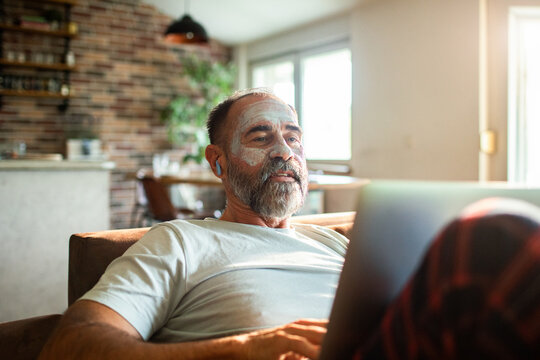 Mature man relaxing with face mask using laptop at home - Powered by Adobe