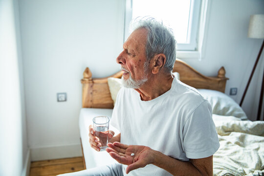 Senior man taking medication with water in bedroom, pensive - Powered by Adobe