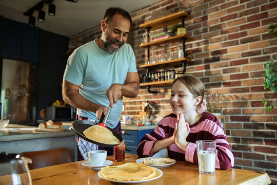 Adult father cooking crepes for happy teen daughter in home kitchen