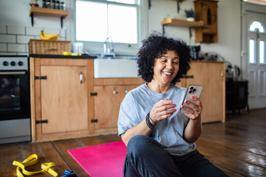 Mature woman smiling at smartphone after workout in home kitchen - Powered by Adobe