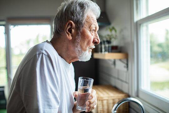 Senior man looking pensive, drinking water by kitchen window - Powered by Adobe