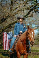 Obraz premium Cowgirl Woman With Braid and Scarf Wearing Cowboy Hat and Denim Jacket Riding Brown Horse With American Flag and Tree in Field in the Background