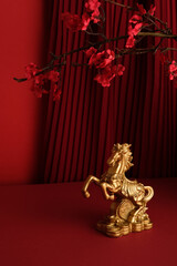 Golden horse statue against a deep red pleated curtain with red blossoms. Happy Chinese New Year 2026 concept.