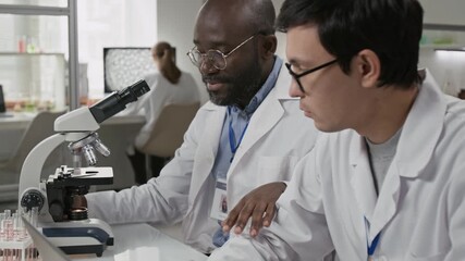 Tilt-up shot of African American scientist asking his colleague to look into microscope while working side-by-side at desk in laboratory - Powered by Adobe