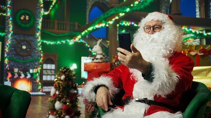 Naklejka premium Upbeat Santa Claus doing videocall with kids using smartphone, bringing joy. Santa Claus entertaining children during phone videoconference, spreading sense of wonder during Christmas, camera A