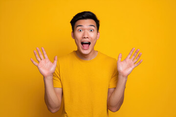Young Asian man in yellow shirt with surprised expression and raised hands against yellow background, energetic joyful emotion and studio portrait