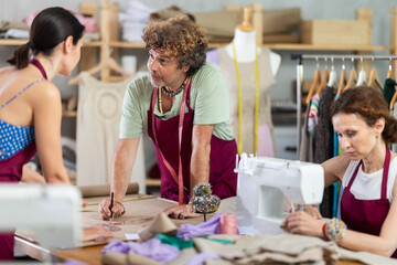Friendly tailor and tailoress drawing on paper and speaking as other working with sewing-machine