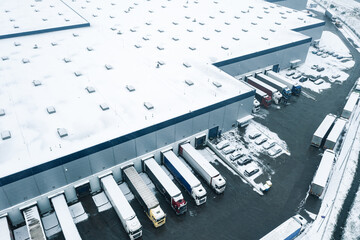 Winter aerial view of a large logistics warehouse in an industrial area with cars and trucks waiting to be unloaded and loaded.