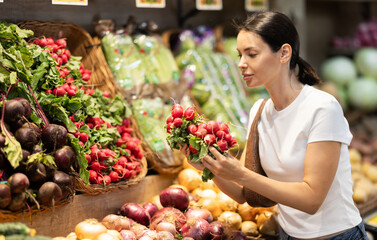 Adult woman buyer choosing fresh radishes in vegetable shop