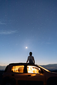 Traveler sitting on a car roof, gazing at the night sky, lost in deep thoughts and spiritual connection beneath the stars.