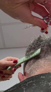 Vertical close-up of barber cutting gray-haired man hair with scissors and comb from low angle. Scene place inside barbershop or beauty salon, highlighting professional grooming and men&rsquo;s hairstyling.