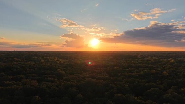A wide cinematic aerial drone shot capturing a glowing golden-hour sunset over a dense forest landscape. The sun lights up the sky with warm color, dramatic cloud formations, and deep atmospheric rays