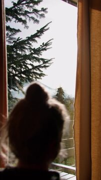 Vertical 9x16 shot of a woman opening curtains to the morning sun, seen from behind, with autumn scenery outside and warm light streaming beautifully through the window.