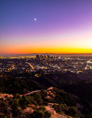 Los Angeles cityscape at dusk from a hillside, featuring a vibrant twilight sky with purple, orange, and yellow hues, sparkling city lights, and a crescent moon.