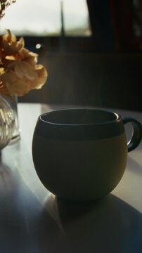 Quiet moment of a tea cup set on a kitchen table as steam rises gently, beautifully backlit by soft autumn morning sunlight, creating a serene, cozy atmosphere.