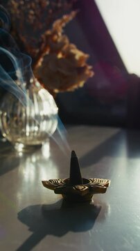 9x16 vertical close-up of smoke rising from a burning incense cone by a sunlit window on an autumn morning, with flowers on the table creating a serene, meditative, calming mood.