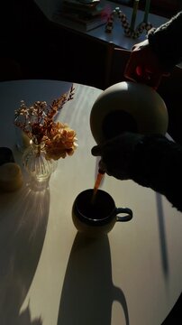 Top-down vertical shot of a relaxing morning tea ceremony as a woman&rsquo;s hands pour tea from a ceramic pot, softly lit by the sun, with steam rising and glowing in the backlight.