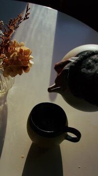 Top-down vertical shot of a relaxing morning tea ceremony as a woman&rsquo;s hands pour tea from a ceramic pot, softly lit by the sun, with steam rising and glowing in the backlight.
