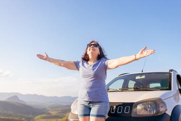 Joyful mature Brazilian woman enjoying freedom on a sunny road trip, standing with open arms beside her car and celebrating independence after 60