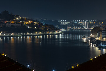 Fototapeta premium Night view of the Douro River in Porto, Portugal, featuring illuminated riverside buildings and the Arrábida Bridge reflected on the water.