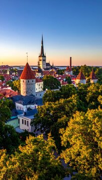 Panoramic view of Tallinn Old Town at sunset, Estonia, with historic buildings and lush green trees under a vibrant sky.