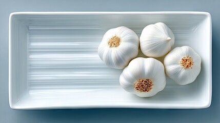 Four whole garlic bulbs are neatly arranged in a white, rectangular dish. The dish has a subtle ribbed texture on the inside. The background is a solid, muted b