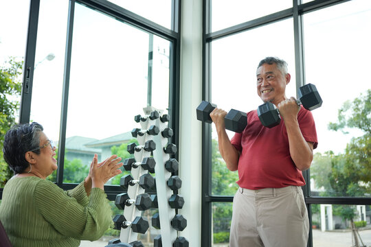 Senior Man Lifting Dumbbells With Supportive Partner, Elderly Couple Exercising Together At Home Gym, Active Senior Training With Dumbbells While Woman Cheers