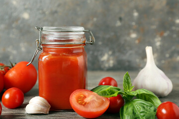Jar of tasty ketchup with garlic, fresh tomatoes and basil leaves on wooden table against grunge grey wall