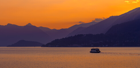 Golden hour ferry crossing tranquil Lake Como Italy at sunset under glowing orange sky and misty Italian Alps mountains serene travel landscape romantic escape dreamy evening over still water panorama