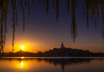 Golden sunset over Beihai Park lake in Beijing China with White Dagoba temple silhouette framed by willow branches reflection tranquil travel landscape zen evening serenity and peaceful golden water